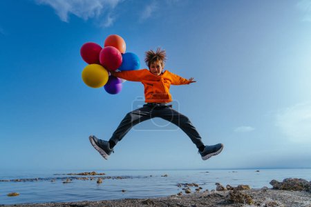 Photo for Cheerful boy jumping with balloons on lake shore on summer day - Royalty Free Image