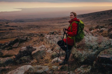 Téléchargez les photos : Jeune femme randonneuse avec des bâtons de trekking reposant contre un rocher, lumière douce du coucher du soleil - en image libre de droit