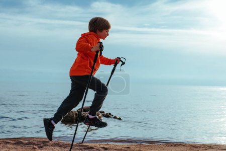 Téléchargez les photos : Joyeux garçon randonneur saute avec des bâtons de trekking sur la rive du lac - en image libre de droit