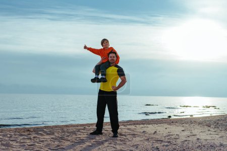 Foto de Niño feliz se sienta en el hombro de su padre y muestra los pulgares hacia arriba en el fondo de la orilla del lago - Imagen libre de derechos