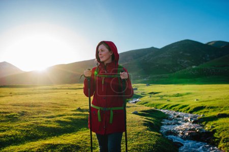 Photo for Young woman tourist standing by stream in picturesque mountain valley on a sunny day - Royalty Free Image