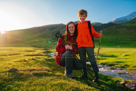 Téléchargez les photos : Jeune touriste avec son fils assis au bord du ruisseau dans la vallée pittoresque de montagne par une journée ensoleillée - en image libre de droit