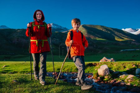 Téléchargez les photos : Jeune touriste avec son fils se promène le long du ruisseau dans la vallée pittoresque de montagne par une journée ensoleillée - en image libre de droit