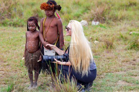 Foto de INDONESIA, PAPUA NEW GUINEA, WAMENA, IRIAN JAYA, 20 AUGUST 2018: A woman with long hair and a camera in her hand sits next to and holds the hand of the children from the papuan tribe in Wamena, Papua - Imagen libre de derechos
