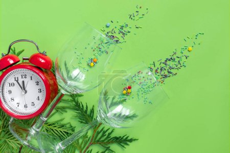 Foto de Reloj despertador con ramas de árbol y copas de champán sobre fondo de papel verde. 5 minutos antes del Año Nuevo. Tarjeta de felicitación creativa - Imagen libre de derechos