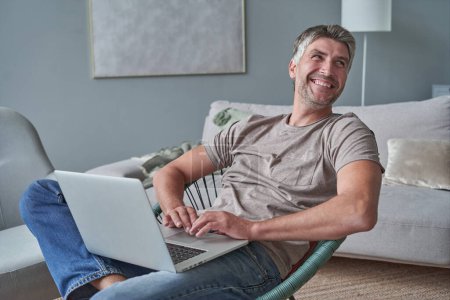 Téléchargez les photos : Jeune homme souriant attrayant navigue sur son ordinateur portable - en image libre de droit