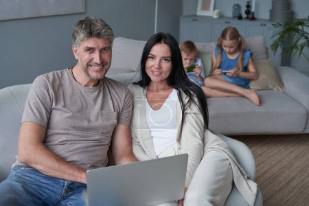 Téléchargez les photos : Fermer parents heureux à l'aide d'un ordinateur portable assis sur le canapé à la maison. Mère et père souriants regardant l'écran d'ordinateur portable - en image libre de droit