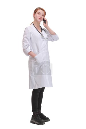 Téléchargez les photos : Portrait de jeune femme médecin debout sur fond blanc isolé tout en faisant un appel téléphonique sur son smartphone et en regardant la caméra - en image libre de droit