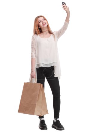 Foto de Mujer joven y bonita feliz con bolsas de compras en ropa vaquera hace foto autorretrato en el teléfono inteligente sobre fondo blanco - Imagen libre de derechos