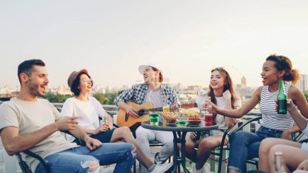Téléchargez les photos : Le gars créatif en vêtements décontractés joue de la guitare et ses amis multi-ethnique chantent en tenant des bouteilles avec de la bière et des boissons gazeuses assis en cercle à table sur le toit
. - en image libre de droit