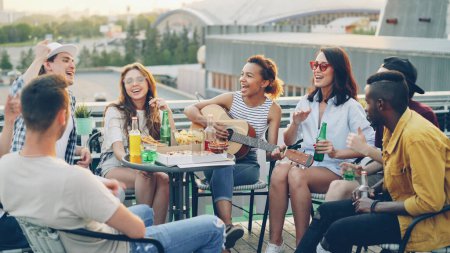 Photo for Pretty African American girl is playing the guitar while her happy friends multiethnic group are singing and moving hands enjoying party on rooftop. Table with food and drinks is visible. - Royalty Free Image