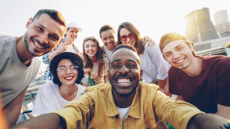 Foto de Punto de vista foto de jóvenes alegres grupo multiétnico tomando selfie y sosteniendo la cámara, hombres y mujeres están mirando a la cámara, sonriendo y posando con bebidas en la fiesta en la azotea
. - Imagen libre de derechos