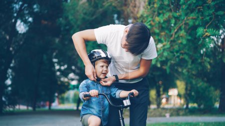 Téléchargez les photos : Le père attentionné met un casque de sécurité sur la tête de ses petits fils puis enseigne à un garçon heureux à faire du vélo dans un beau parc - en image libre de droit