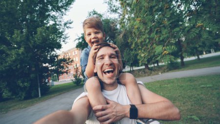 Foto de POV de padre feliz llevando a su hijo sonriente en el cuello y haciendo selfie video en la cámara del teléfono inteligente durante el paseo en el parque - Imagen libre de derechos