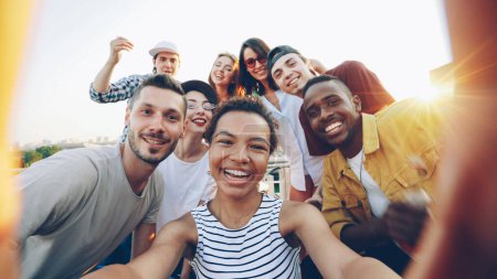 Téléchargez les photos : Point de vue prise de vue de jolie fille afro-américaine tenant la caméra et prenant selfie avec des amis heureux à la fête sur le toit. Hommes et femmes regardent la caméra, posant et riant
. - en image libre de droit