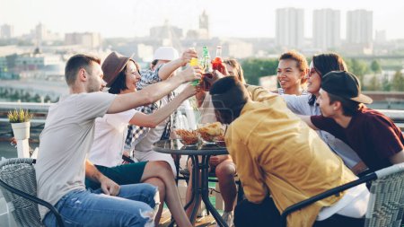Photo for Multiethnic group of friends is talking, toasting then clanging bottles and glasses and drinking beer and soft drinks at table on roof. Beautiful view of big city is in background. - Royalty Free Image