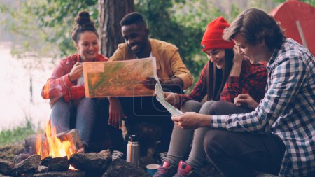 Photo for Male and female friends tourists are sitting around fire and studying maps during hike in forest in summer. Young people are wearing casual clothing. - Royalty Free Image