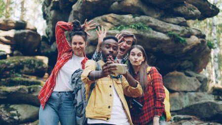 Foto de Feliz hombre afroamericano turista se está tomando selfie con amigos en el bosque cerca de enormes rocas utilizando el teléfono inteligente, la gente está haciendo caras divertidas y mostrando gestos de mano fresca
. - Imagen libre de derechos