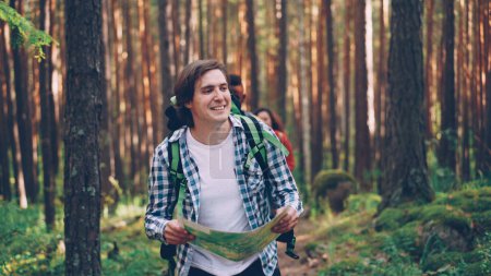 Photo for Handsome young guy is holding map and walking in forest while his friends are following him along trail in wood. Travelling, summertime and orientation concept. - Royalty Free Image