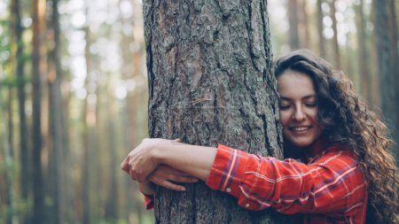 Téléchargez les photos : Belle jeune femme aux cheveux bouclés portant une chemise lumineuse étreint arbre profitant de la nature et souriant avec les yeux fermés. Gens joyeux, concept de loisirs et de bonheur
. - en image libre de droit