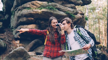 Photo for Cheerful couple is standing in forest, man is studying paper map, girl is looking through binoculars then showing interesting place to her friend and laughing. - Royalty Free Image
