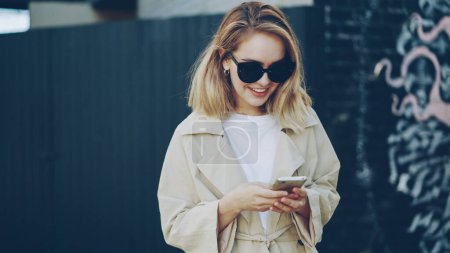 Téléchargez les photos : Joyeux jeune femme utilise smartphone textos son ami et souriant debout à l'extérieur dans la ville moderne avec la peinture graffiti en arrière-plan. Les gens, les émotions et la technologie concept
. - en image libre de droit