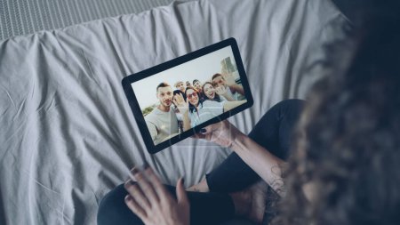 Téléchargez les photos : Fille aux cheveux bruns parle avec des amis en ligne à l'aide d'une tablette en regardant l'affichage, geste et main agitant. Technologie moderne, chat avec des amis et concept joyeux millennials
. - en image libre de droit