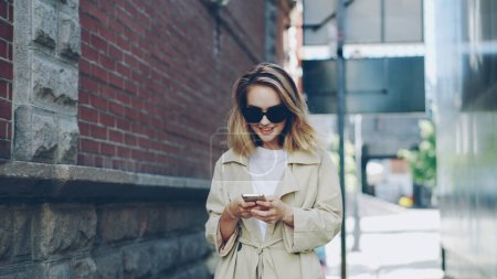 Photo for Pretty blond girl is using modern smartphone touching screen walking in city and smiling enjoying device. Young woman is wearing fashionable clothing and trendy sun glasses. - Royalty Free Image