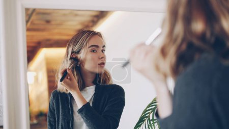Foto de Chica bonita joven está terminando maquillaje tocando la cara y el cuello con cepillo y luego mirando en el espejo grande y sonriendo disfrutando de un aspecto hermoso. Enfoque en la reflexión espejo
. - Imagen libre de derechos