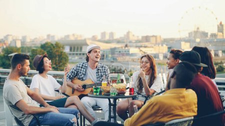 Téléchargez les photos : Joyeux musicien masculin joue de la guitare avec ses amis chantant et riant assis sur le toit à table. Divertissement, instruments de musique et concept de loisirs
. - en image libre de droit