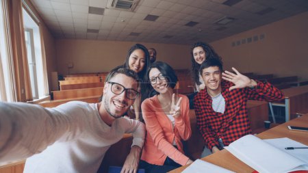 Foto de Punto de vista plano de grupo multiétnico de amigos estudiantes tomando selfie en el aula mirando a la cámara, posando con gestos de mano y sonriendo. Tecnología moderna y concepto de los millennials
. - Imagen libre de derechos