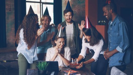 Photo for Cheerful girl office worker is working at desk while her coworkers are bringing birthday cake and putting party hat on her, excited young woman is clapping hands. - Royalty Free Image