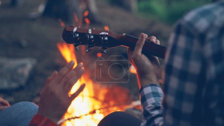 Photo for Close-up shot of male hands playing the guitar and female hands moving in dance with burning campfire in background. Camping, music and adventurous people concept. - Royalty Free Image