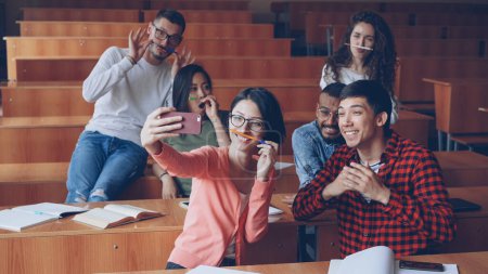 Photo for Playful young people students are taking funny selfe using pens and pencils as moustache, posing and showing hand gestures thumbs-up and v-sign sitting in classroom. - Royalty Free Image