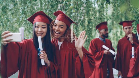 Foto de Dos chicas atractivas compañeros estudiantes se están tomando selfie con teléfono inteligente en el día de la graduación con diplomas, las mujeres jóvenes están sonriendo, posando, haciendo gestos de manos y caras divertidas
. - Imagen libre de derechos