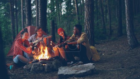 Photo for Male tourist is playing the guitar sitting near campfire with friends singing and having fun, young people are holding sticks with marshmallow above flame. Food, music and fun concept. - Royalty Free Image