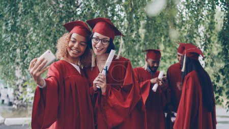 Foto de Las mujeres bonitas graduadas están tomando selfie con el pergamino del diploma usando el teléfono inteligente, las mujeres jóvenes están posando con otros estudiantes en vestidos moviéndose y hablando en segundo plano
. - Imagen libre de derechos