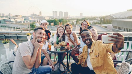 Photo for Young African American man is holding smartphone and taking selfie with his friends multi-ethnic group holding bottles and glasses, posing and enjoying rooftop party. - Royalty Free Image