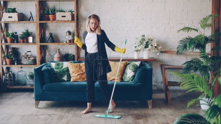 Photo for Attractive girl in headphones is listening to music and dancing wih mop during domestic work, she is mopping floor at home and having fun. Women, joy and modern houses concept. - Royalty Free Image