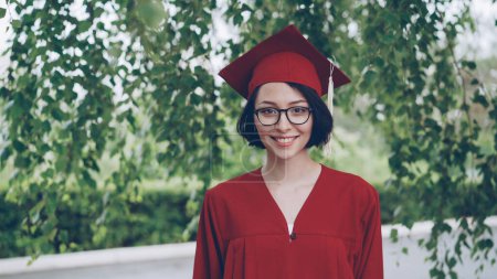 Téléchargez les photos : Portrait d'une jeune femme joyeuse diplômée en robe rouge et en mortier souriant et regardant la caméra debout sous l'arbre sur le campus. Concept jeunesse et éducation
. - en image libre de droit