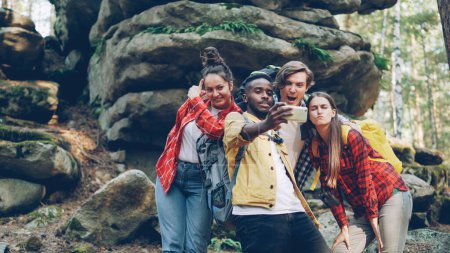 Foto de Grupo multirracial de amigos turistas modernos se están tomando selfie en el bosque con rocas en el fondo usando teléfonos inteligentes, hombres y mujeres están posando y mostrando gestos de la mano
. - Imagen libre de derechos