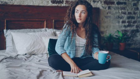Photo for Pretty girl is reading book sitting on bed at home and holding cup with drink enjoying free time and resting. Young people, hobby and modern interior concept. - Royalty Free Image