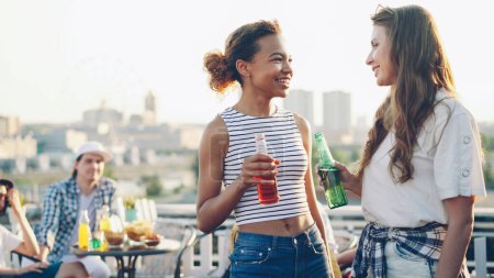 Photo for Pretty African American girl is talking to her cheerful Caucasian friend and drinking cocktail holding bottle during open-air party on rooftop. Friendship, fun and communication concept. - Royalty Free Image