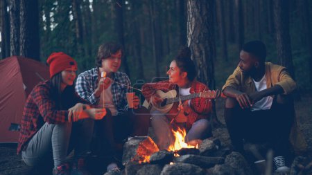 Photo for Pretty young woman is playing the guitar while her friends are singing funny songs and laughing cooking marshmallow on fire and clapping hands. Music, nature and fun concept. - Royalty Free Image