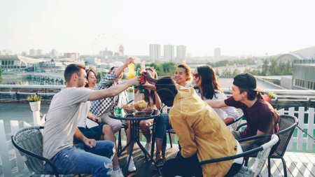 Photo for Attractive young men and women are toasting and clinking glasses and bottles then drinking beer and cocktails and laughing during party on rooftop in modern city. - Royalty Free Image
