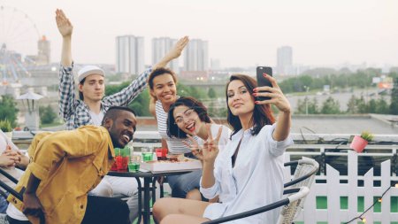 Photo for Pretty young woman is holding smartphone and taking selfie with emotional friends sitting at table with food and drinks on rooftop. Beautiful city is in background. - Royalty Free Image