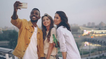 Stylish African American guy is taking selfie with cute Caucasian girls at open-air party on rooftop, young man is using modern smartphone, woman is holding beer bottle.