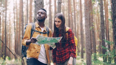 Photo for Multiracial couple is looking at map, talking during getaway in wild forest in summer on sunny day. Trees, grass and backpacks are visible. - Royalty Free Image