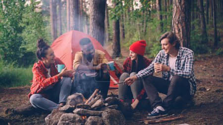 Téléchargez les photos : Groupe multiethnique d'amis les jeunes filles et les gars sont assis dans la forêt autour du feu avec des verres cliquetis et souriant, la fumée monte. - en image libre de droit
