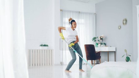 Photo for Cheerful African American girl in gloves is mopping floor, listening to music with wireless headphones and dancing enjoying rhythm cleaning her nice modern apartment. - Royalty Free Image